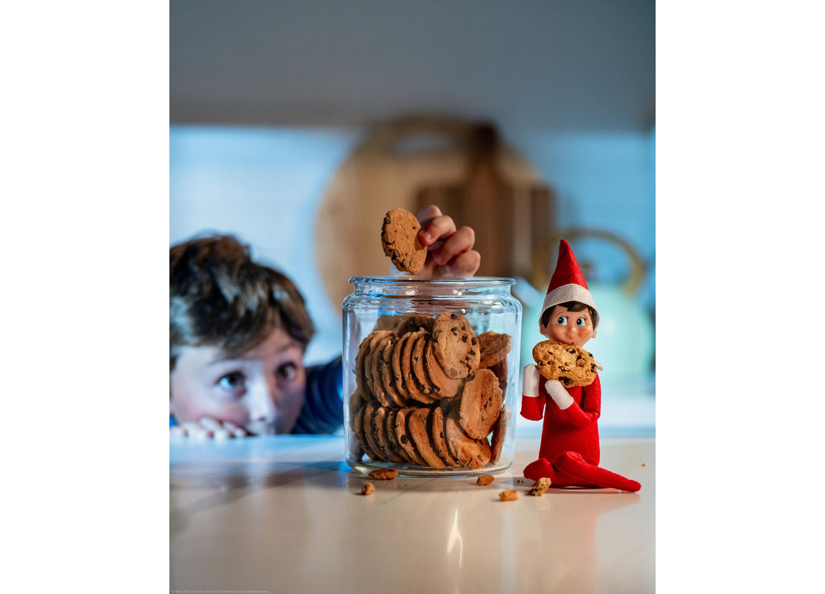 Elf on the Shelf Scout Elf Boy figure with red outfit and pointed hat, holding chocolate chip cookie next to glass ja...