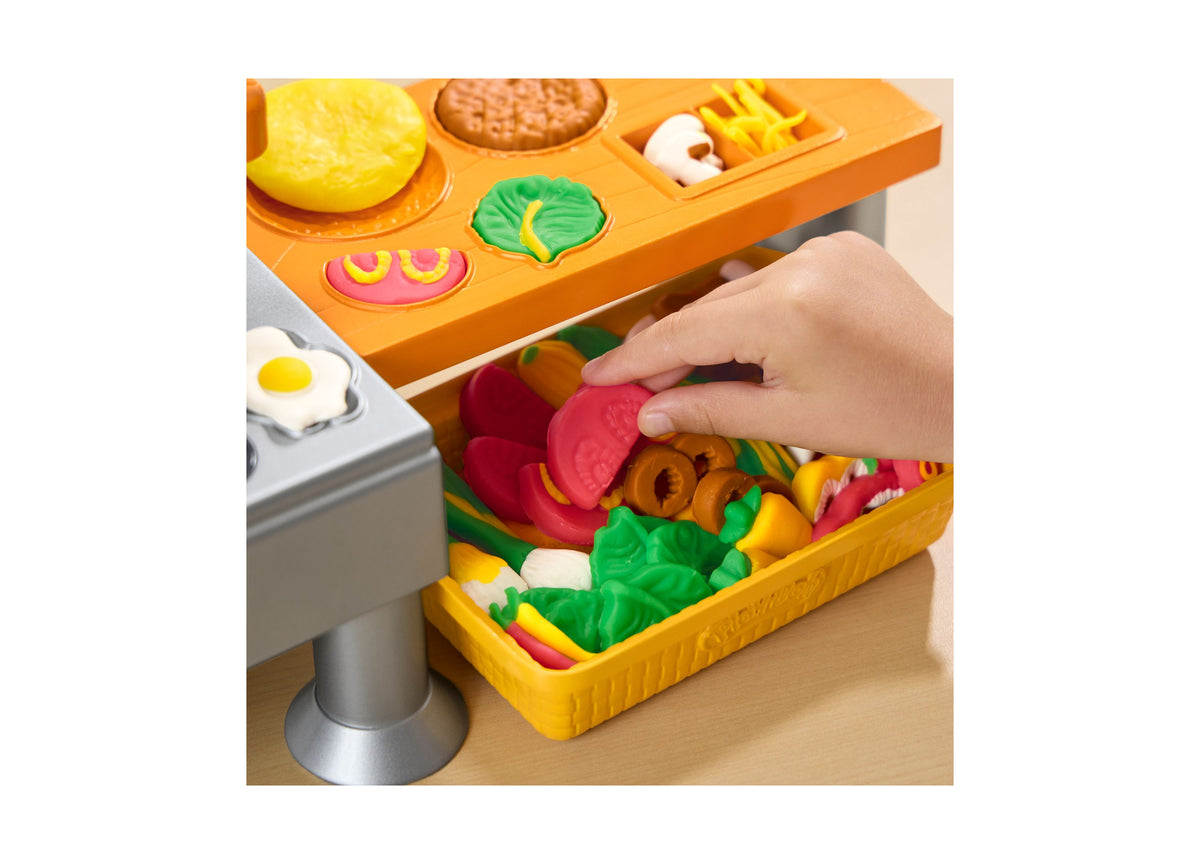 Play-Doh Super Sizzlin Kitchen Playset with child's hand selecting red tomato-shaped food from yellow storage drawer
