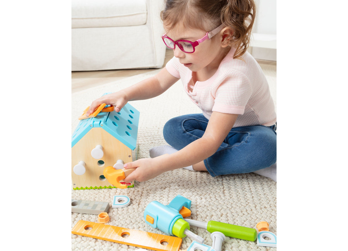Child in pink shirt and glasses building toy house with colorful blue, yellow, and green components from Melissa and ...