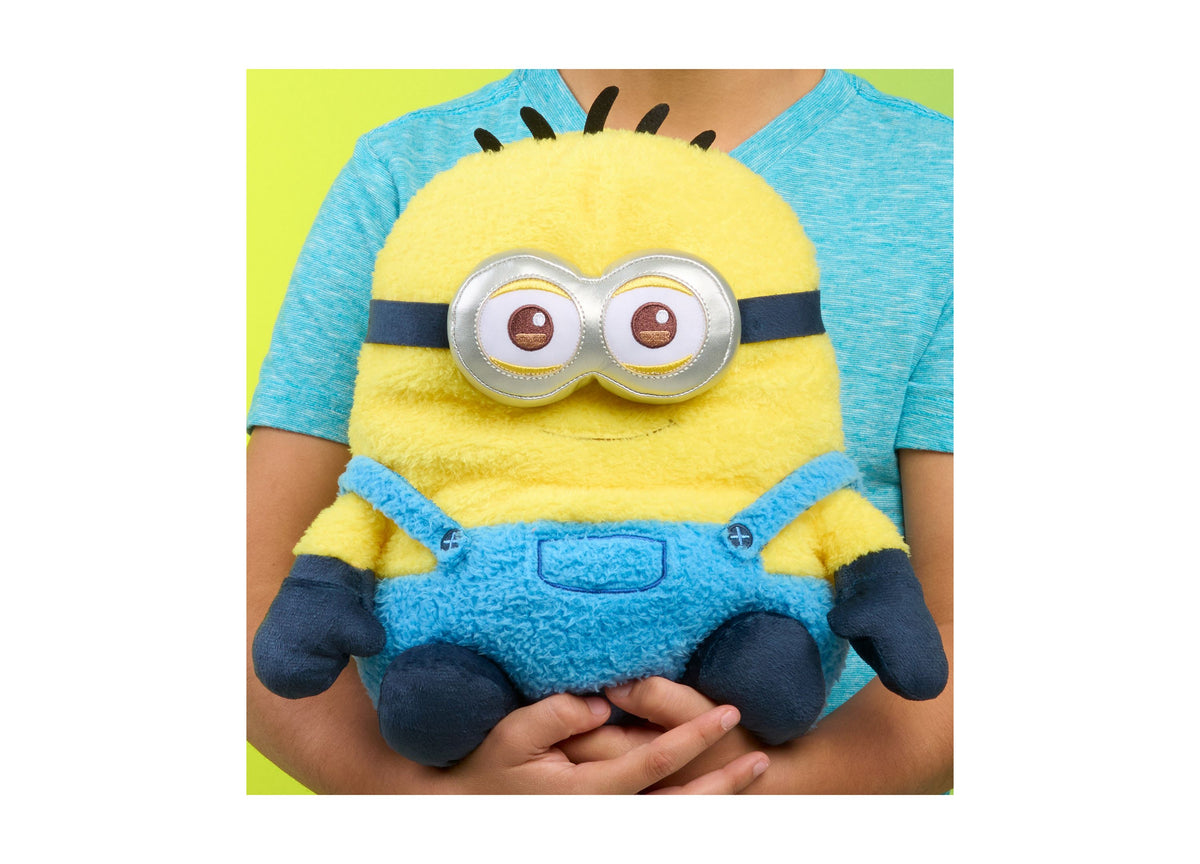 Minions Ed 10" Weighted Comfeez Plush toy held in hands, featuring yellow body, blue overalls, large goggle eyes, and...