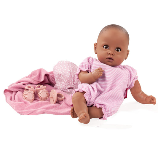Baby doll with brown skin and sleeping eyes wearing pink ribbed dress and bonnet, posed on pink blanket with small shoes