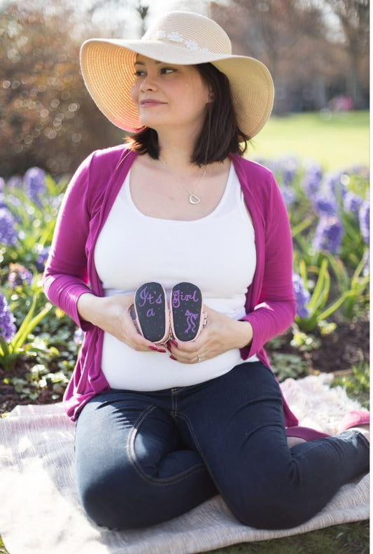 Pregnant woman on model holding chalkboard baby moccasins in garden setting, wearing wide-brimmed straw hat and magenta cardigan