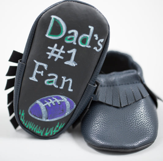 Gray moccasin with black fringe trim and chalkboard sole reading "Dad's #1 Fan" with football illustration, shown from sole-up angle
