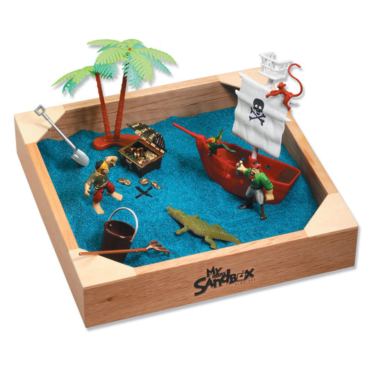 Wooden sandbox with blue sand containing pirate-themed figurines, red ship, palm tree, treasure chest, skull flag, crocodile, shovel, and bucket