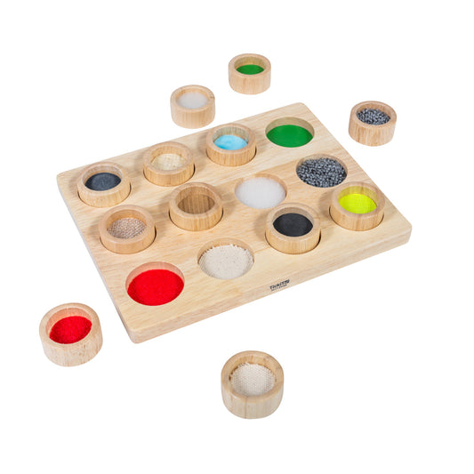 Wooden touch and match board with 16 removable lidded cups containing different textured materials in various colors including red felt, blue beads, green gel,