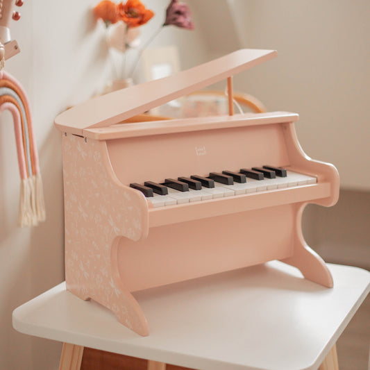 Pink wooden toy piano with black and white keys, lid propped open, displayed on white surface with decorative flowers and rainbow decor in background